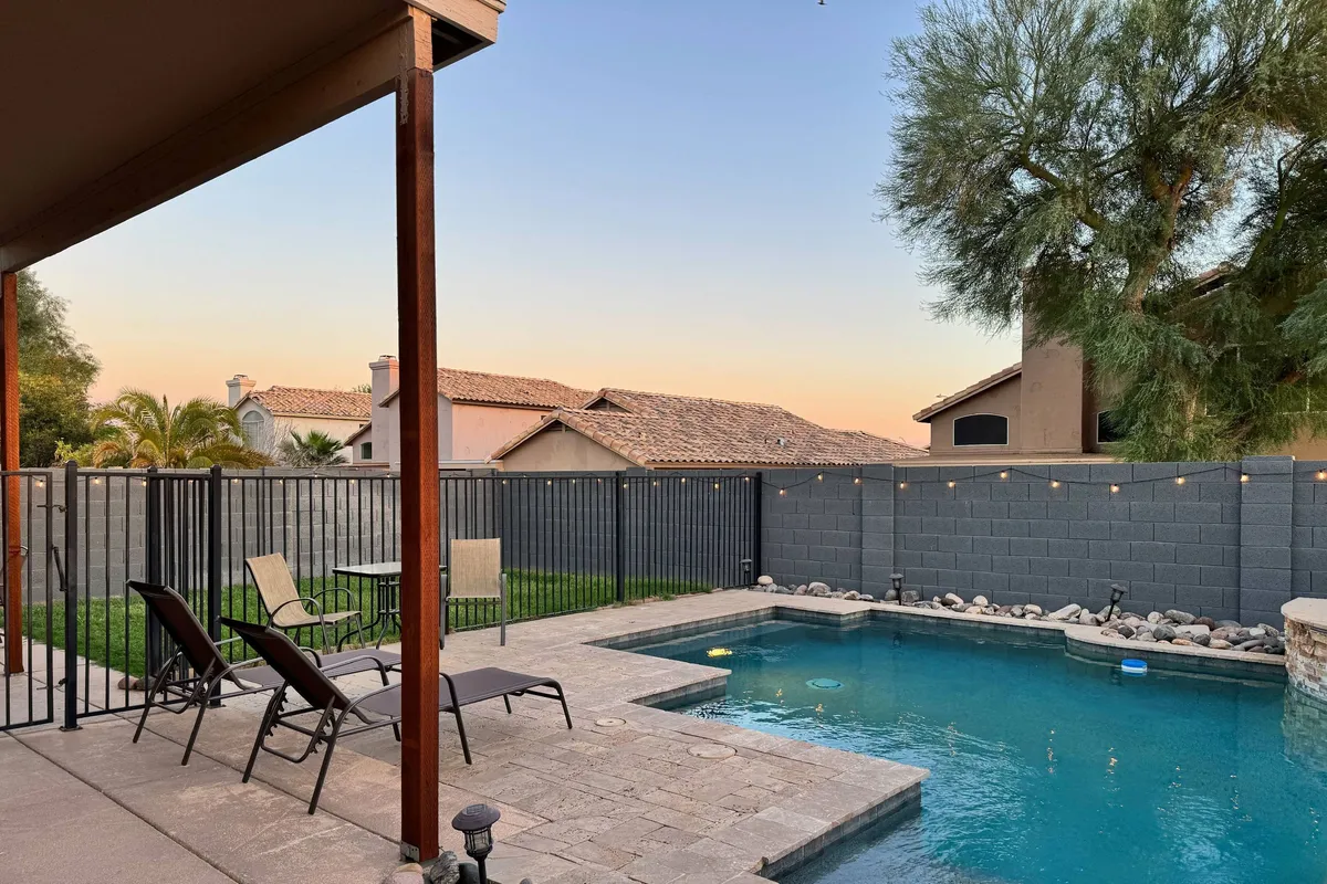 A serene backyard scene featuring a sparkling swimming pool with a waterfall feature, surrounded by multiple lounge chairs. An adjacent outdoor dining area is visible, positioned to soak up the Arizona sun, inviting both relaxation and al fresco meals. The pool area is fully gated, providing safety for families and peace of mind for guests.