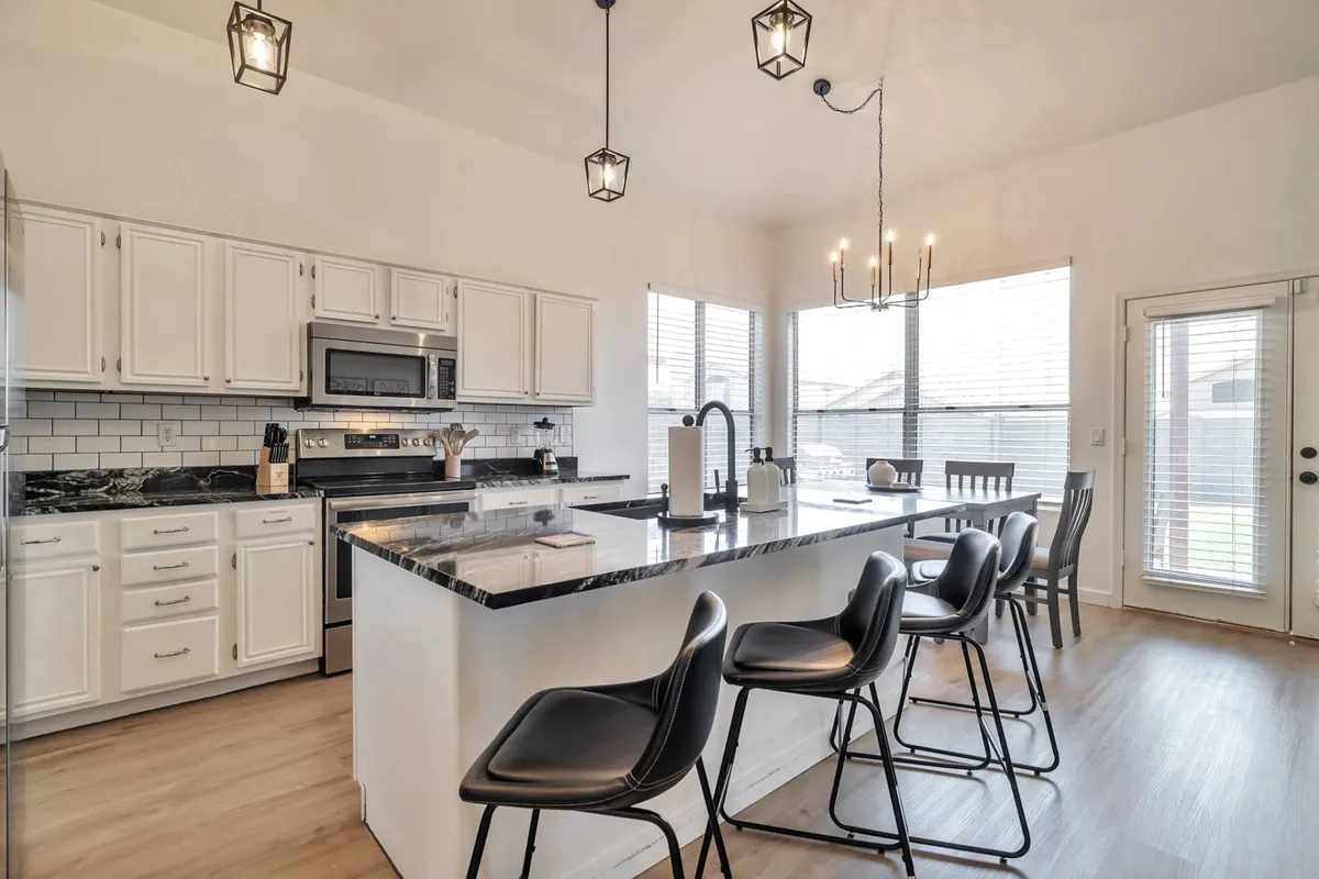 This image showcases a bright, contemporary kitchen featuring a spacious island bar with comfortable seating, modern pendant lighting, stainless steel appliances, and crisp white cabinetry. Large windows let in natural light, creating a welcoming atmosphere perfect for both cooking and gathering.