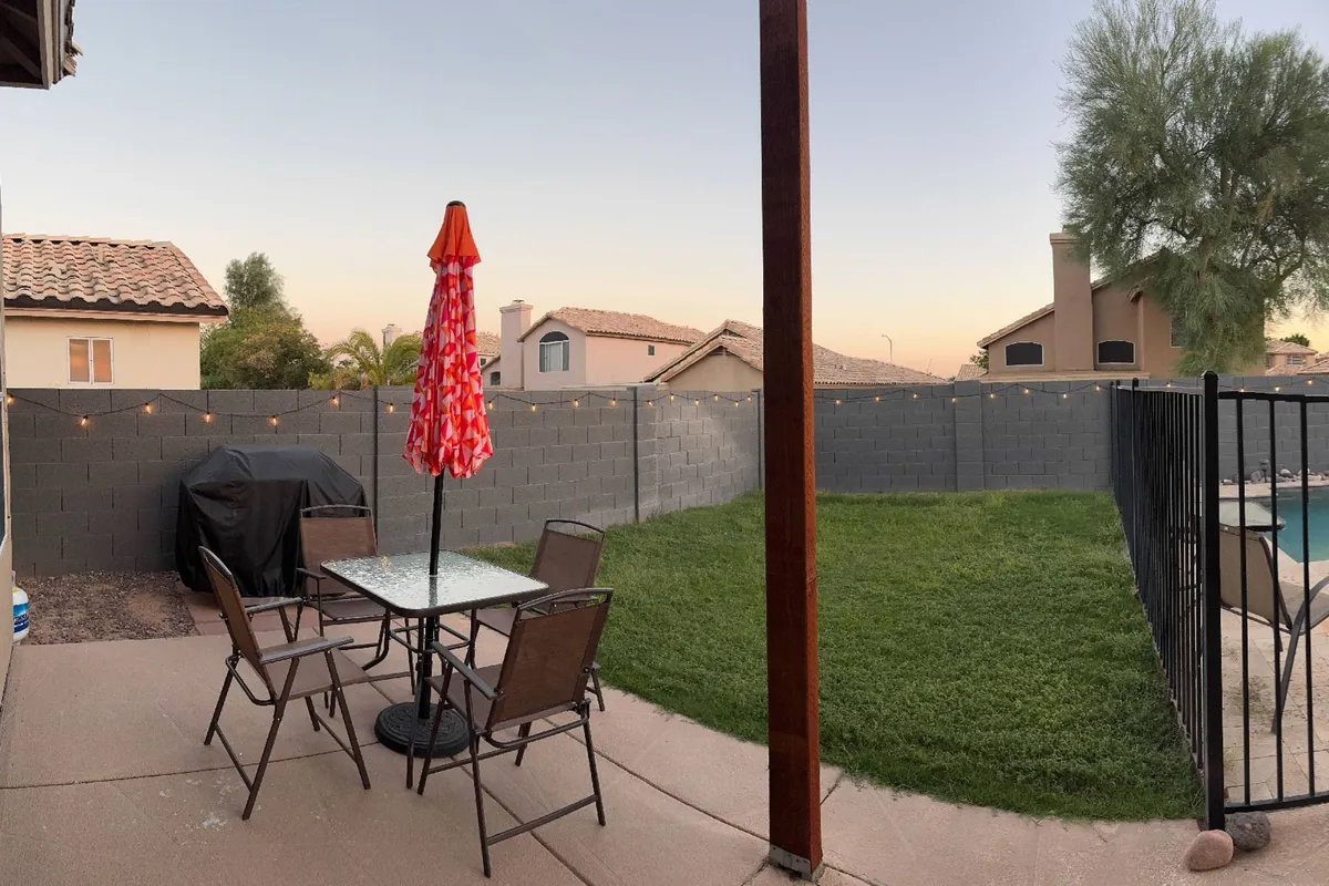 An inviting outdoor gathering space featuring a glass table shaded by a red umbrella, surrounded by comfortable sitting chairs and a lush green lawn. The fenced yard is strung with lights, creating a welcoming atmosphere for dining and evening gatherings.