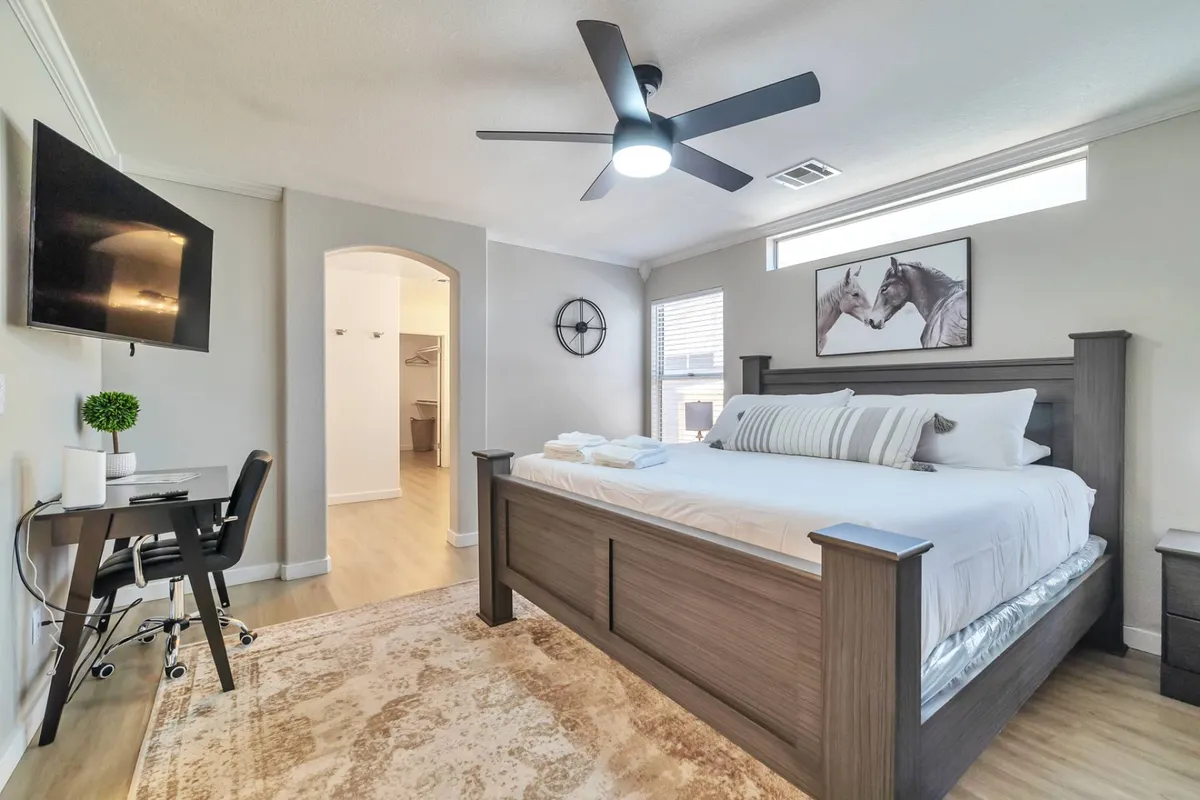 A spacious master bedroom featuring a king-size bed with crisp linens, a wall-mounted smart TV, a dedicated desk and chair for workspace, and tasteful artwork depicting horses. Soft natural light pours in, creating an inviting and restful atmosphere.