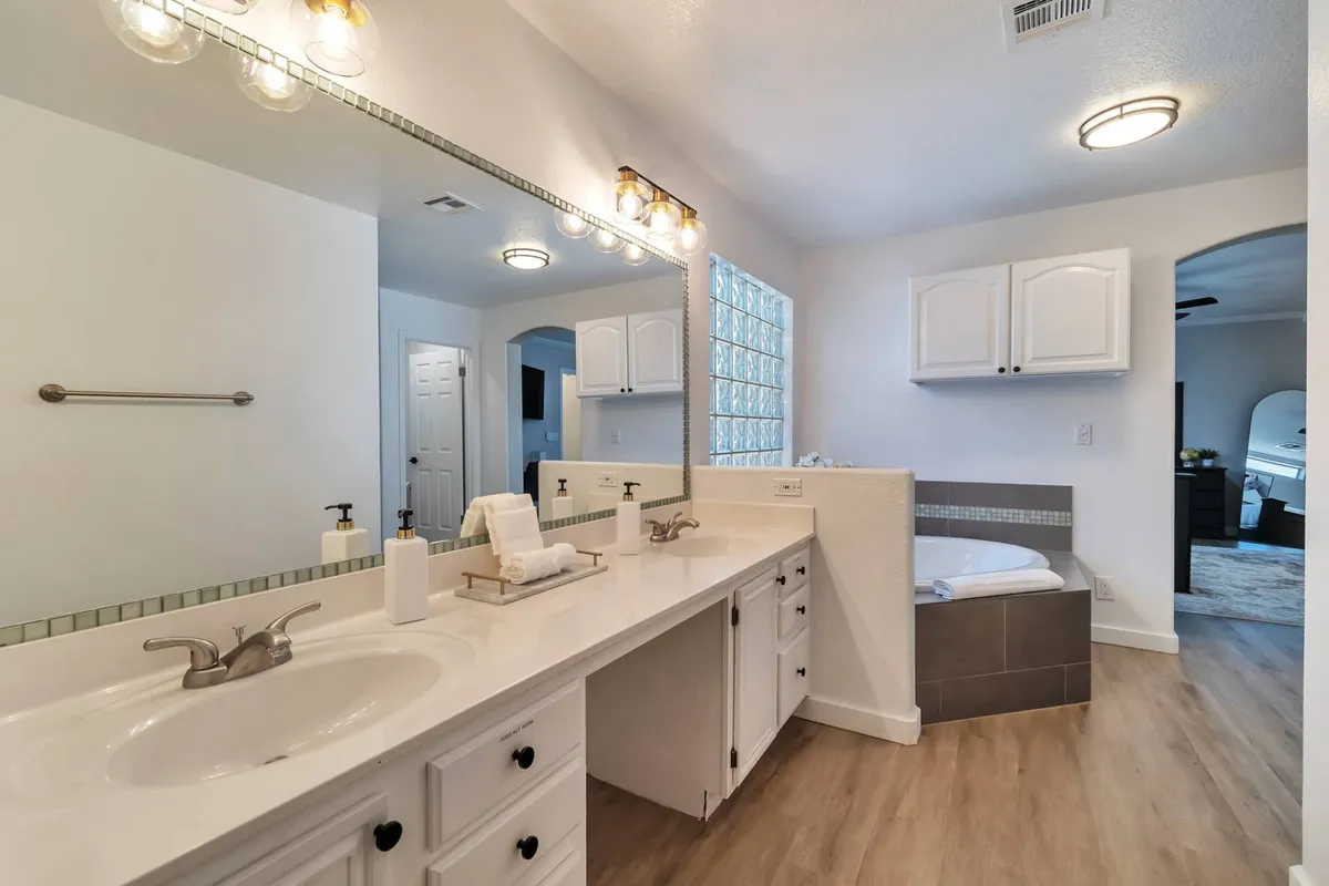 A tranquil bathroom featuring a large soaking tub, double sink vanity, glass block window allowing in natural light, and a spacious mirror. The ambiance is soothing, offering a luxurious spot for self-care and relaxation after a busy day.