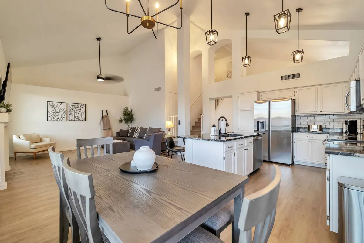 The image showcases a spacious, beautifully lit open-concept area featuring an elegant dining table set for gatherings, a modern kitchen island perfect for cooking and mingling, plush living area seating, and a stylish chandelier that adds a warm, inviting glow. The setup suggests a perfect space for family dinners, festive celebrations, or relaxing after a day out.