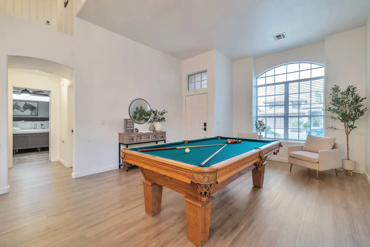 This image showcases a bright, airy living room with a pool table set near a large window, decorative plants, and a seating area that encourages group interaction. The playful, welcoming mood supports various activities from casual lounging to lively pool games.