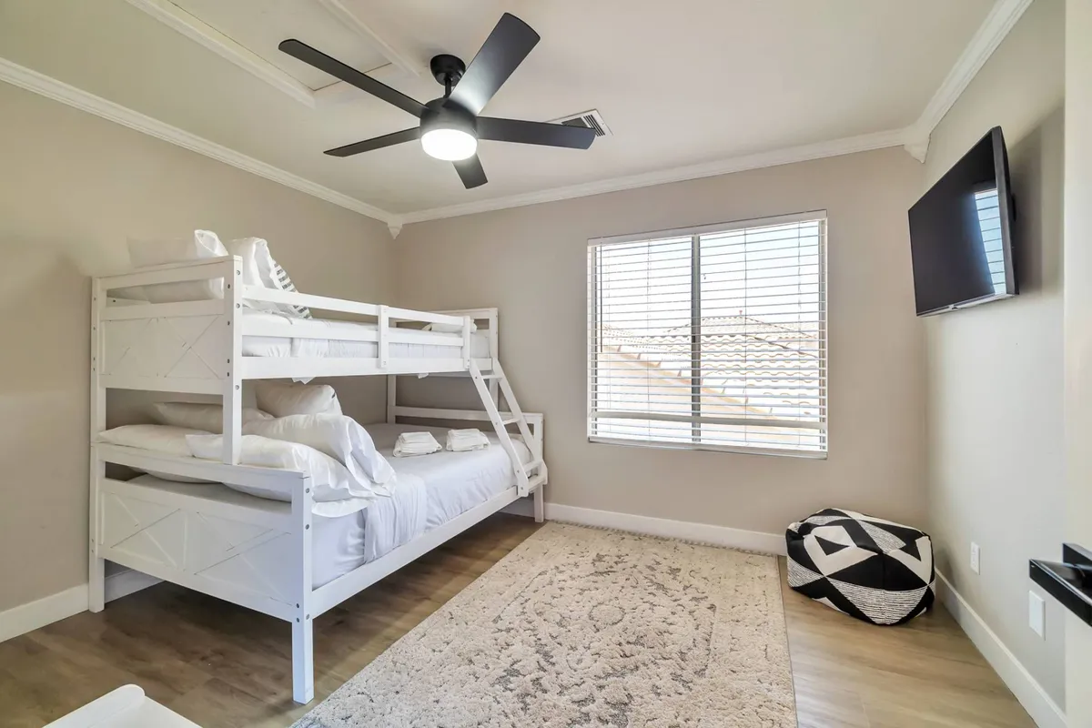 A cozy guest bedroom with a twin/queen bunk bed, colorful rug, ceiling fan, window letting in morning sunlight, and a wall-mounted TV. The space is designed for both comfort and fun, ideal for families or groups with children.