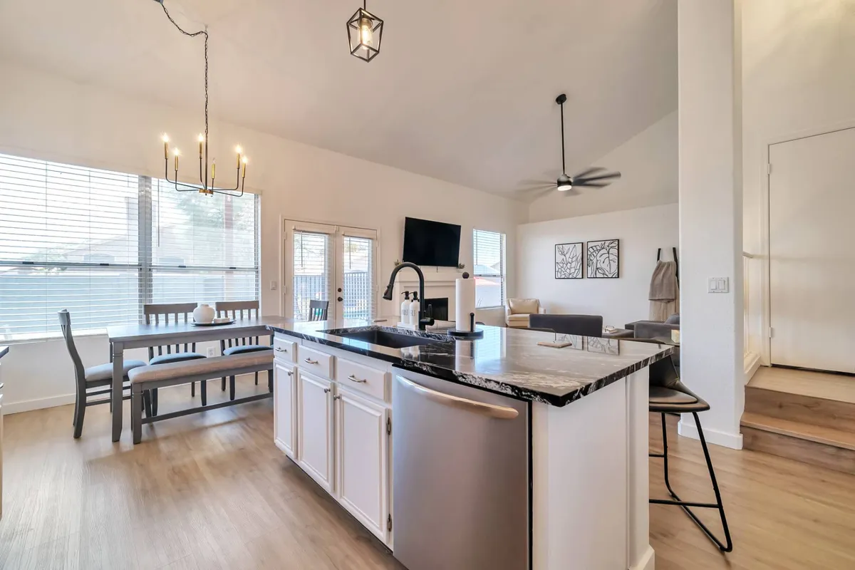 The scene displays a harmonious blend of kitchen, dining, and living spaces connected by a large island, chandelier, and open sightlines. Ample seating and natural light make it clear this is an area designed for communal dining and entertaining.