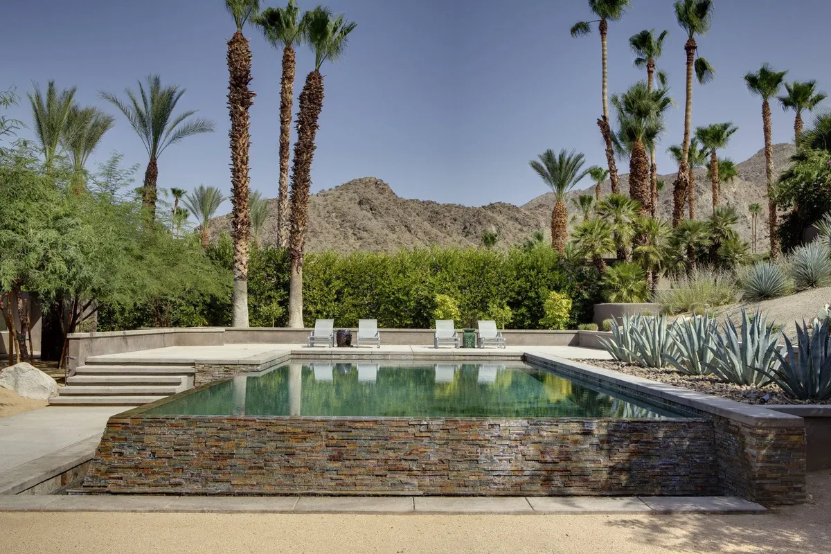 A sparkling swimming pool surrounded by palm trees and lounge chairs, set against a striking mountain backdrop. The pool area is serene, ideal for relaxation or gathering with friends after an active day.