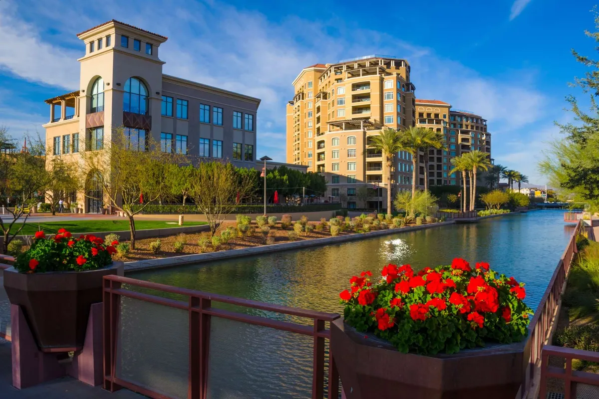 A lively urban scene with modern buildings, blooming flower planters, a canal waterway, and palm trees. The area invites visitors to stroll, dine outdoors, and enjoy the vibrant local community.